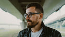 man with a short beard wearing glasses under a freeway overpass.