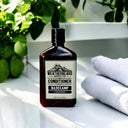 A bottle of Weatherbeard Supply Co. Basecamp conditioner sits on a white surface next to folded white towels and green plants in the background.